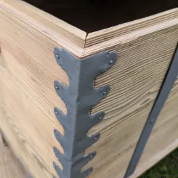 Drewniana skrzynia z metalowymi wzmocnieniami narożników i uchwytem, stojąca na trawie.