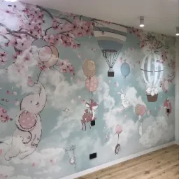 Pokój dziecięcy z fototapetą przedstawiającą zwierzęta unoszące się na balonach na tle chmur i kwitnących drzew wiśni, widoczne drewniane panele podłogowe i oświetlenie sufitowe