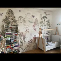 Pokój dziecięcy z fototapetą przedstawiającą leśny krajobraz z jeleniem, grzybami i zwierzętami, białe łóżeczko dziecięce, regał z zabawkami.