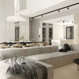 Jasne, przestronne wnętrze: salon przechodzi w jadalnię z długim stołem nakrytym do posiłku, a w tle widoczna kuchnia z marmurowym blatem. Na pierwszym planie szary narożnik z dekoracyjnym węzłem...