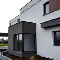 Nowoczesna elewacja domu jednorodzinnego z elementami drewnopodobnymi i dużymi oknami, widoczny fragment ogrodu i ścieżka z płyt.