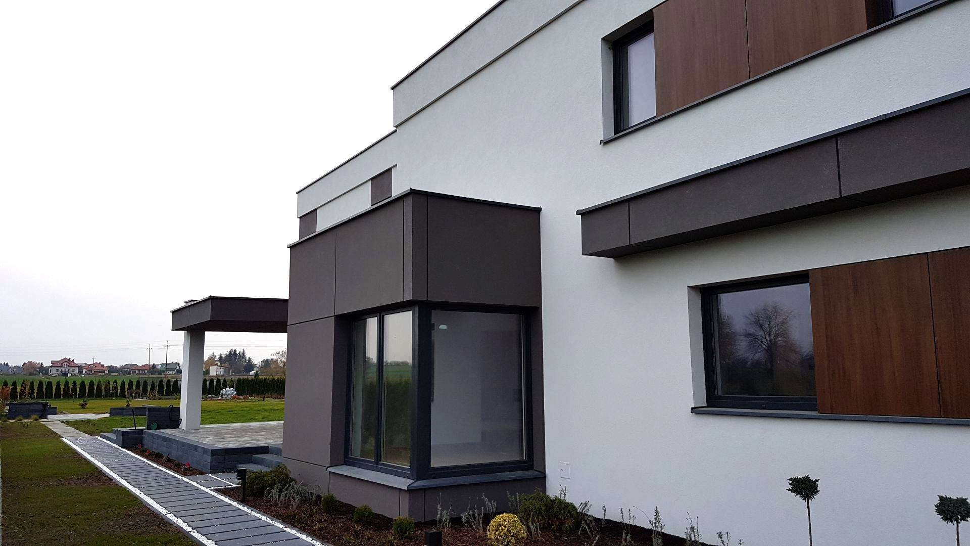 Nowoczesna elewacja domu jednorodzinnego z elementami drewnopodobnymi i dużymi oknami, widoczny fragment ogrodu i ścieżka z płyt.