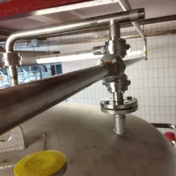 Połączenie rur ze stali nierdzewnej z zaworami i żółtą zaślepką na zbiorniku w pomieszczeniu z białymi płytkami.