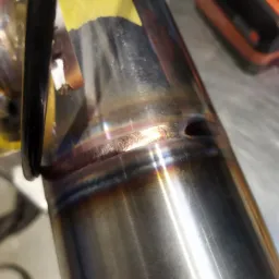 Zbliżenie na spoinę na rurze ze stali nierdzewnej z widocznym otworem, z efektem tęczy na spoinie, obok narzędzia i żółta karteczka z napisem.