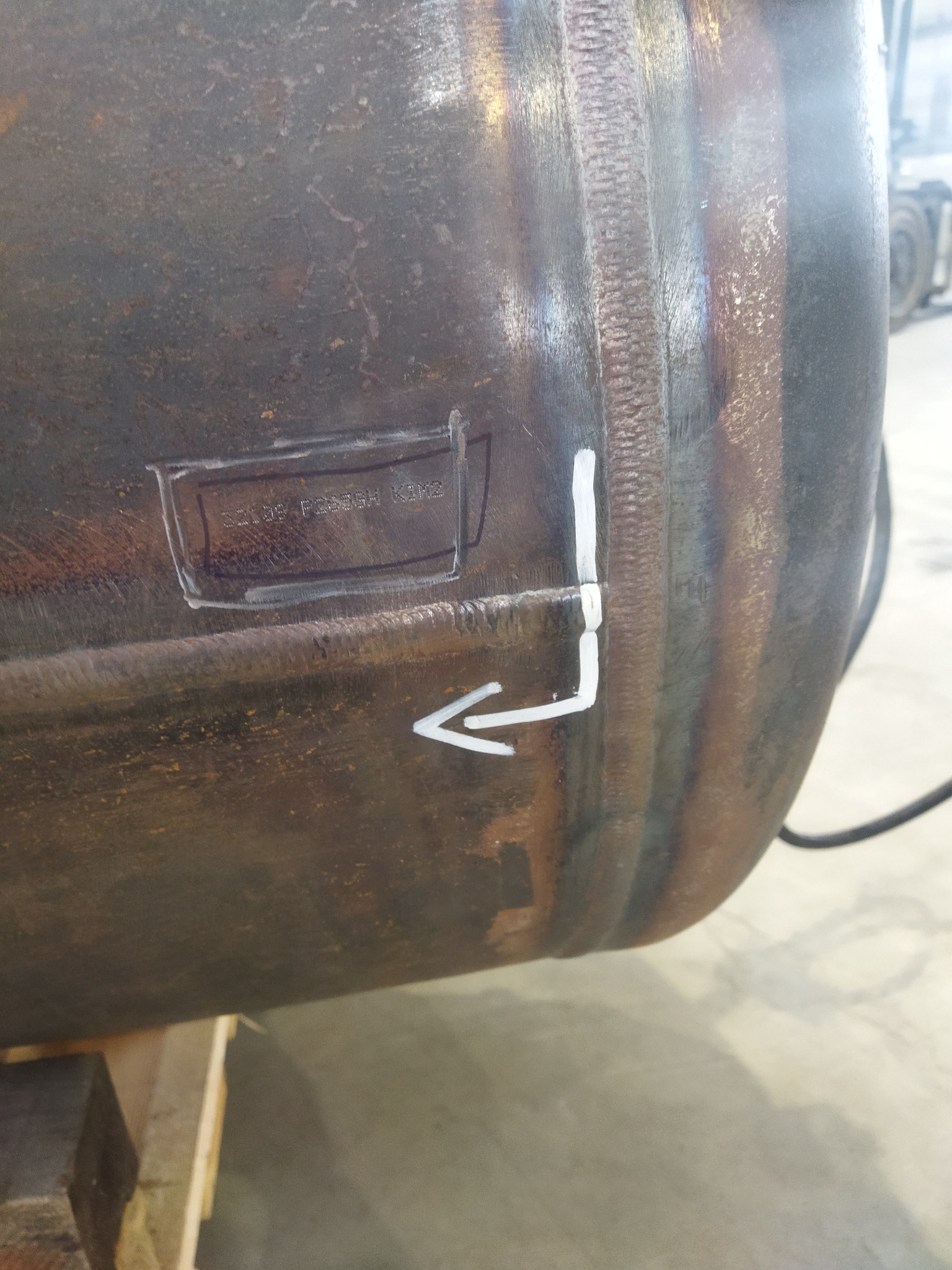 Fragment spawanej, metalowej konstrukcji z zaznaczeniem miejsca spawu białą farbą i napisem w prostokątnej ramce. Widoczne ślady spawania i odbarwienia termiczne.