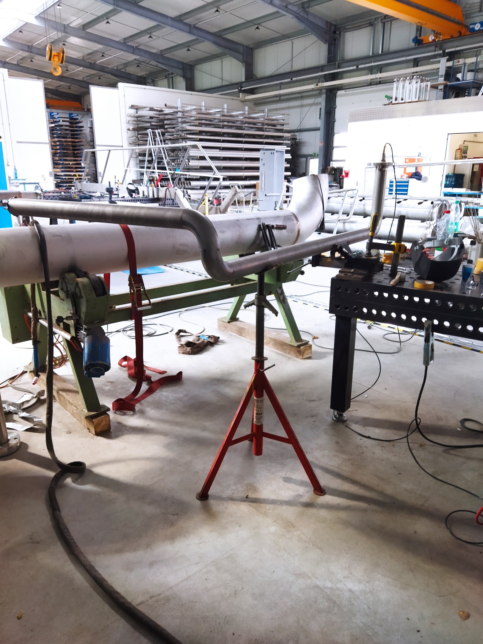 Metalowa rura z zakrzywionym fragmentem, zamocowana na zielonym stojaku i podparta czerwonym trójnogiem, obok stół spawalniczy z maską spawalniczą i narzędziami, w tle hala z regałami pełnymi...