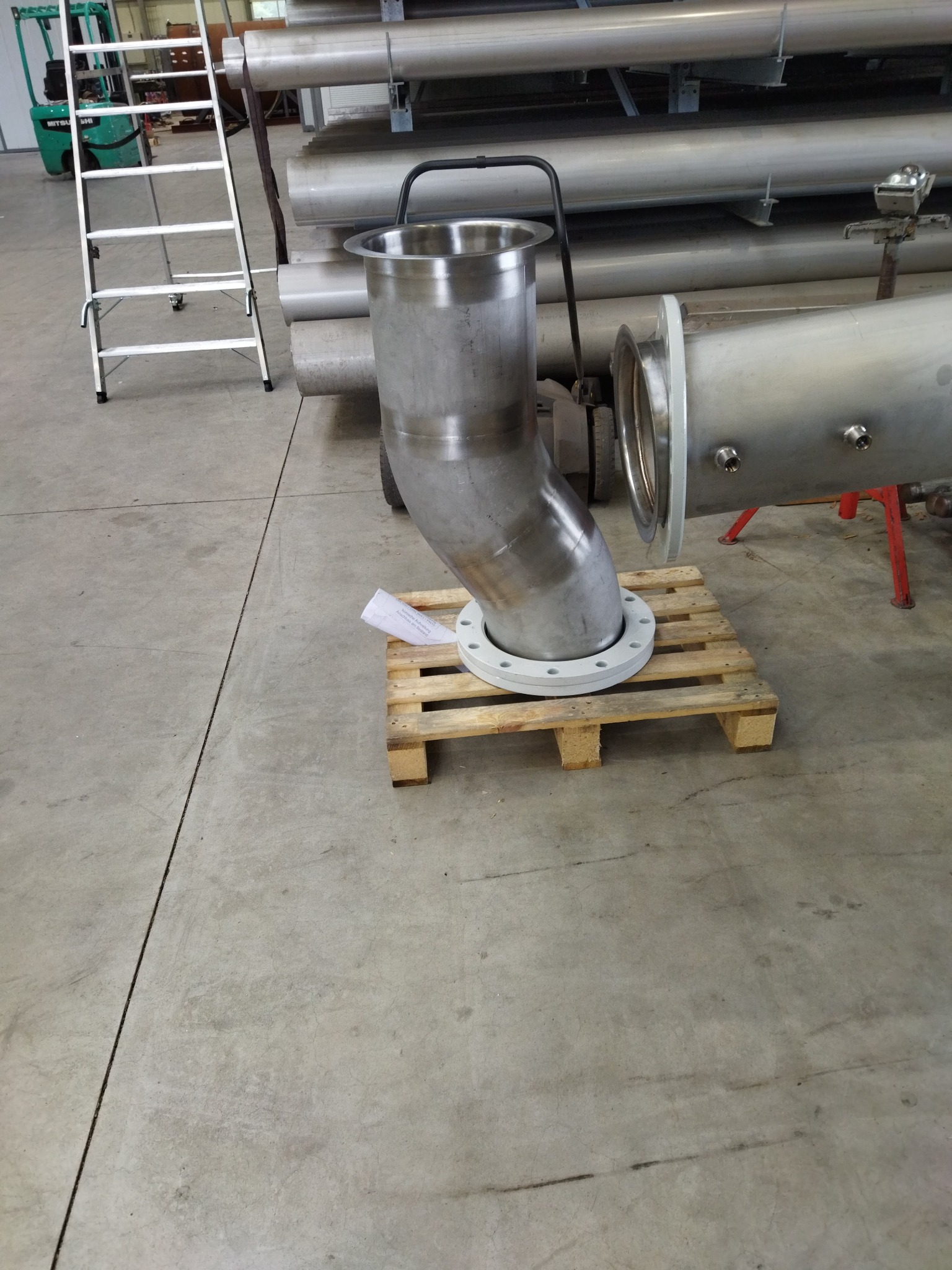 Element rurociągu ze stali nierdzewnej, z kolankiem i kołnierzem, umieszczony na palecie transportowej w hali produkcyjnej, widoczne inne rury i elementy konstrukcyjne w tle.