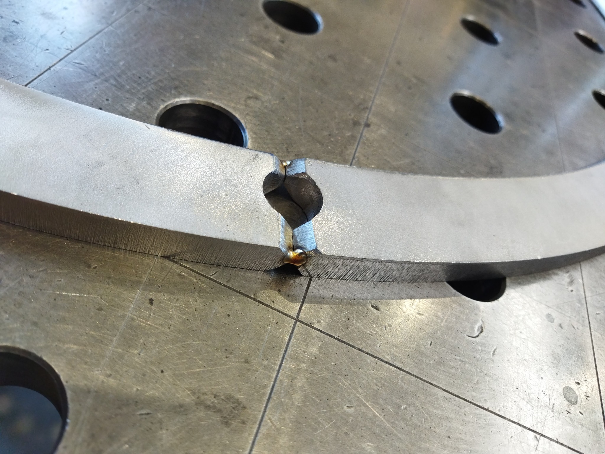 Spoina łącząca fragmenty metalowego pierścienia na perforowanym stole spawalniczym, widoczne ślady obróbki i przypalenia.