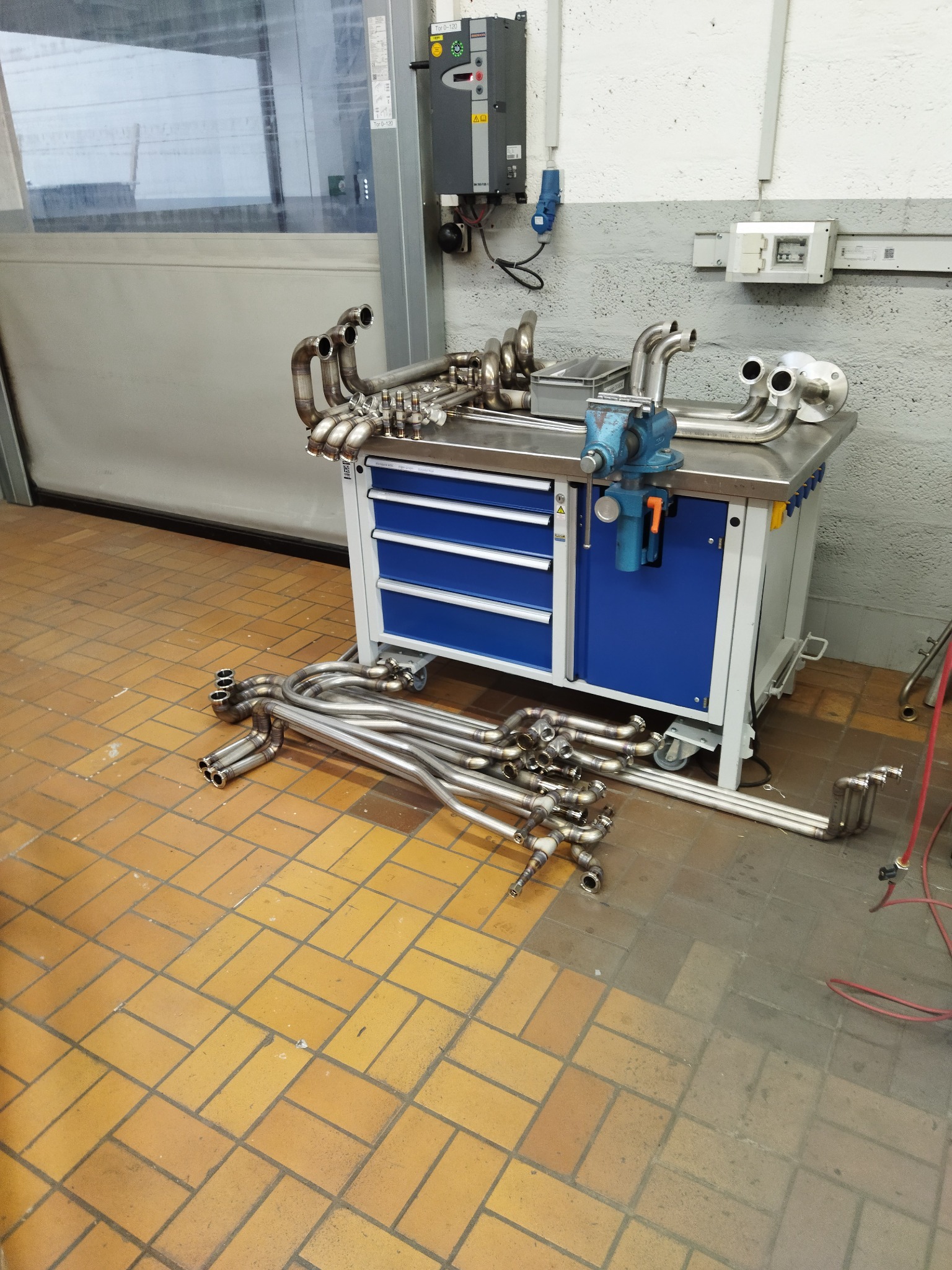 Warsztatowy stół z niepołączonymi, pospawanymi elementami rur ze stali nierdzewnej, na blacie imadło ślusarskie, w tle widoczna brama garażowa.