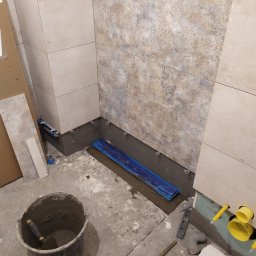 Remonty oraz wykończenia wnętrz - Fragment łazienki w trakcie układania płytek, widoczne narzędzia, wiadro z zaprawą, fragment instalacji wodnej i kanalizacyjnej, oraz płytki o różnej fakturze na ścianach.