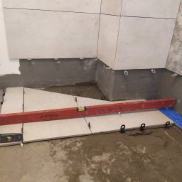 Remonty oraz wykończenia wnętrz - Układanie jasnych płytek podłogowych, z użyciem poziomicy i systemu poziomowania, widoczny świeży klej i elementy montażowe.