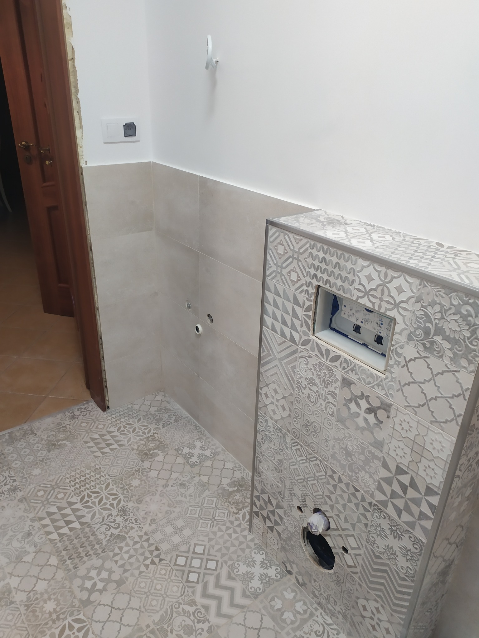 Świeżo wykafelkowana łazienka z wzorzystymi płytkami podłogowymi i na zabudowie stelaża WC, widoczne otwory na przyłącza i niedokończone obramowanie drzwi.