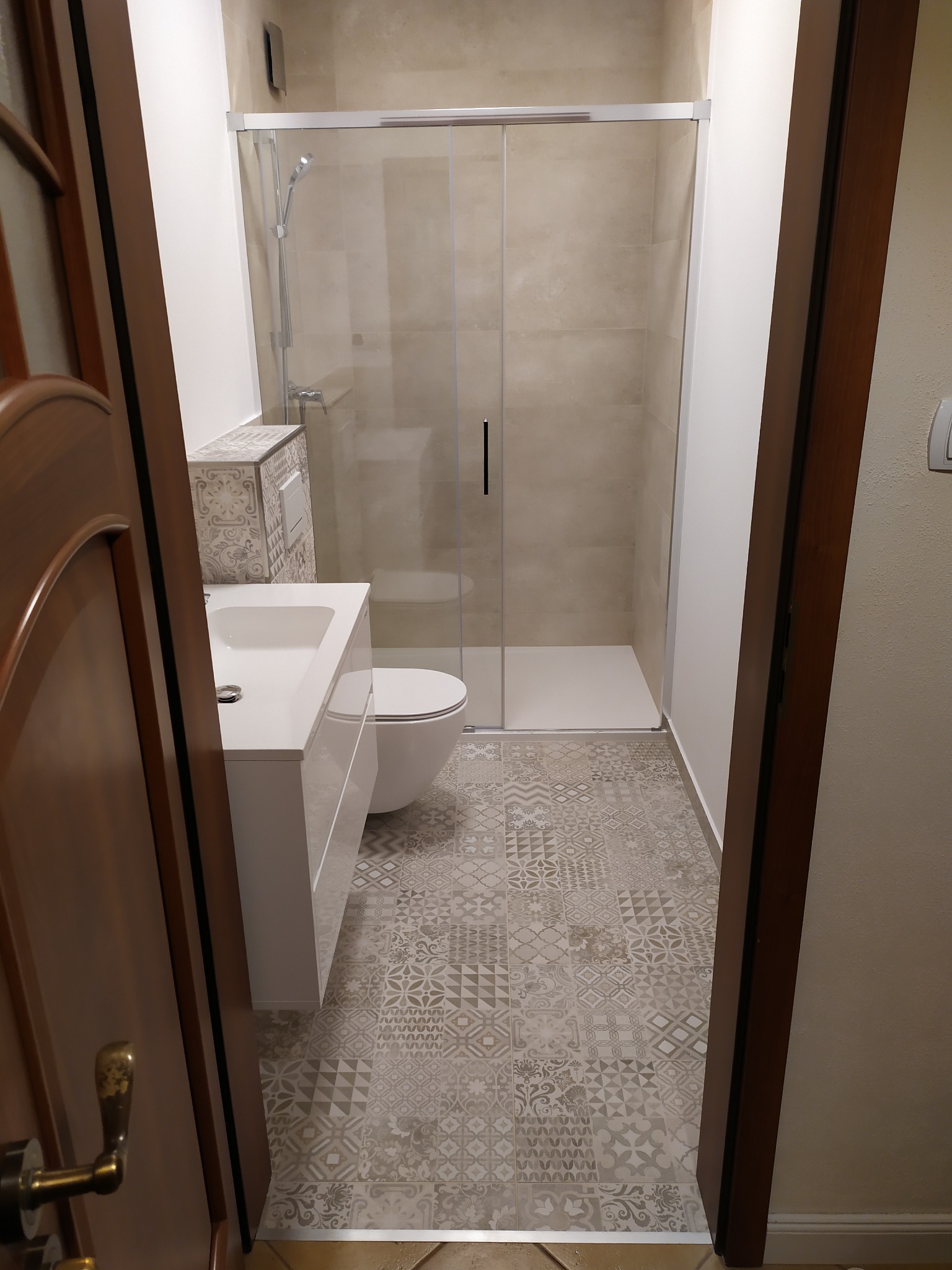 Nowoczesna łazienka z kabiną prysznicową, umywalką z szafką i toaletą, wykończona płytkami podłogowymi w geometryczne wzory, widok zza otwartych drzwi.