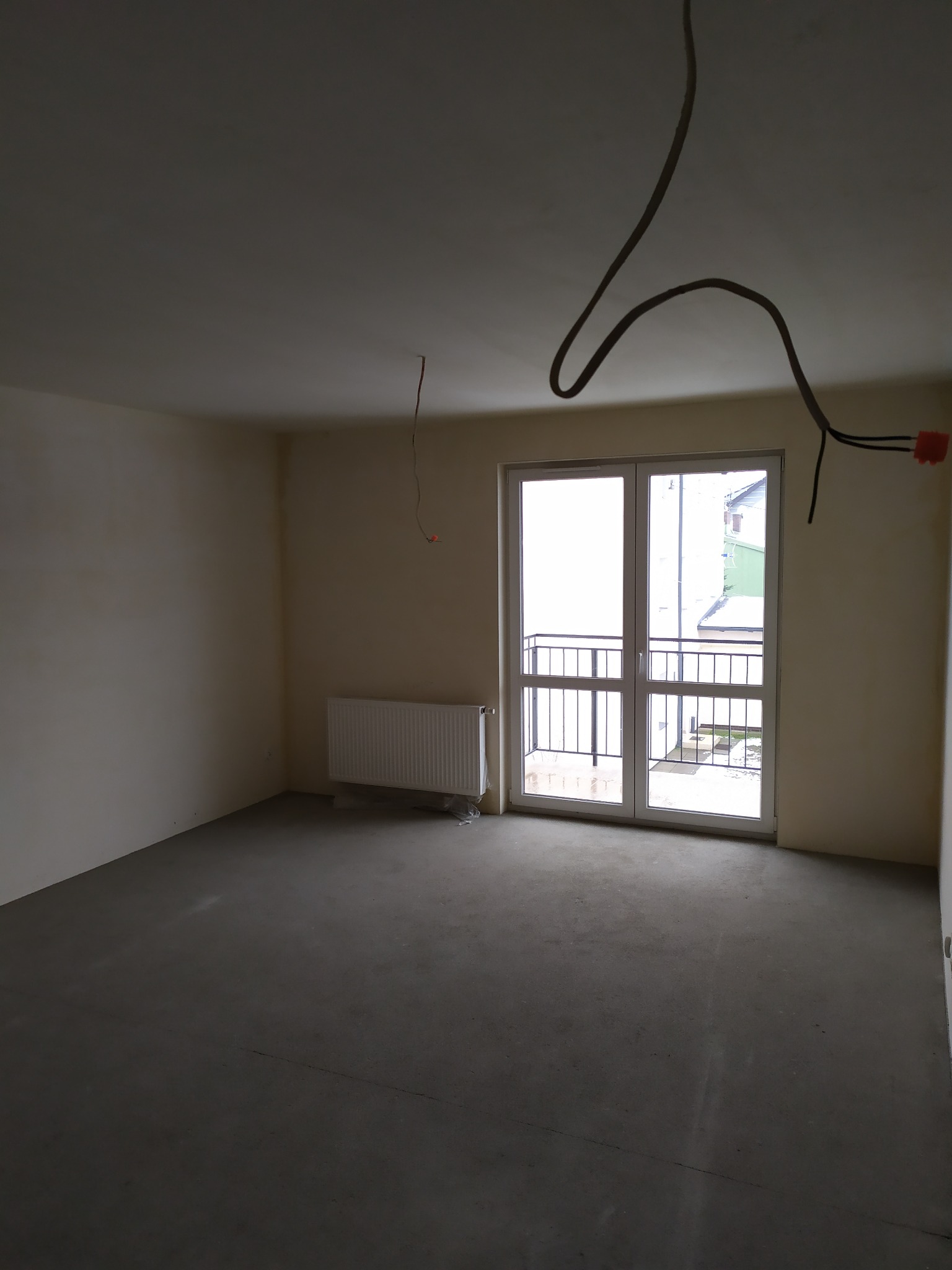 Surowe wnętrze pokoju z wylewką, grzejnikiem i balkonem, ściany przygotowane pod malowanie, widoczne przewody elektryczne.