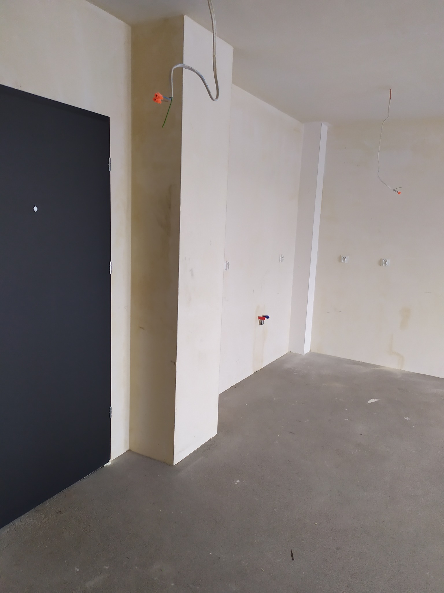 Wnętrze w trakcie prac wykończeniowych: surowe ściany z gładzią, widoczne przewody elektryczne i instalacja wodna, szara wylewka na podłodze, ciemne drzwi wejściowe.