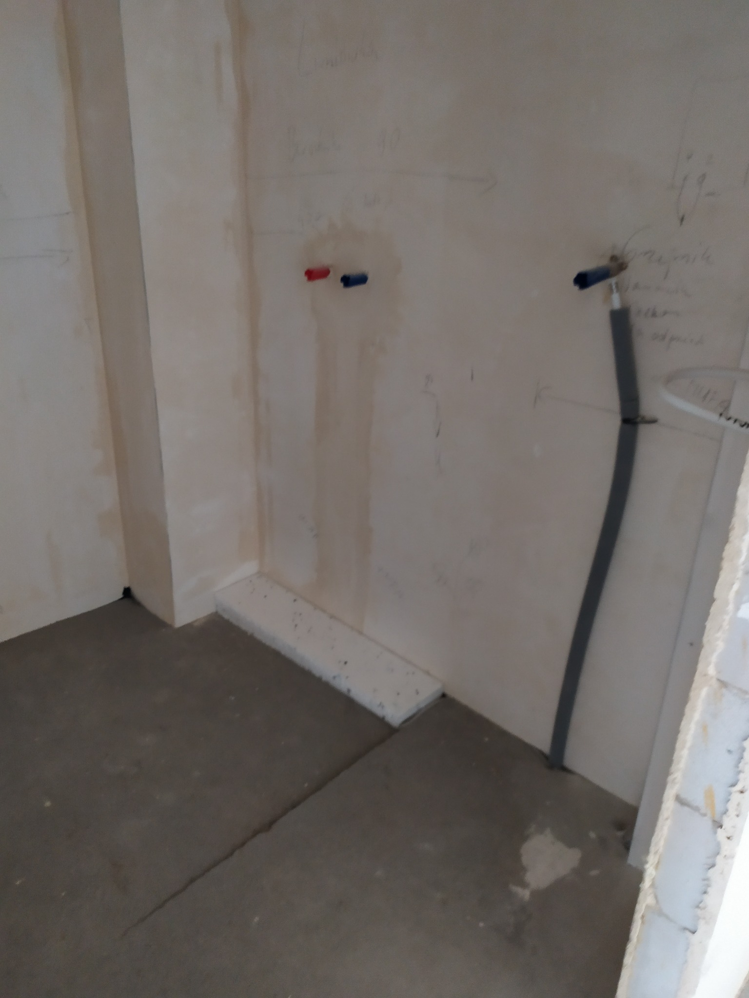 Surowa ściana w trakcie przygotowania do prac wykończeniowych w łazience, widoczne instalacje wodne i elektryczne, oraz odręczne notatki na ścianie, surowa podłoga i fragment ściany z pustaków.