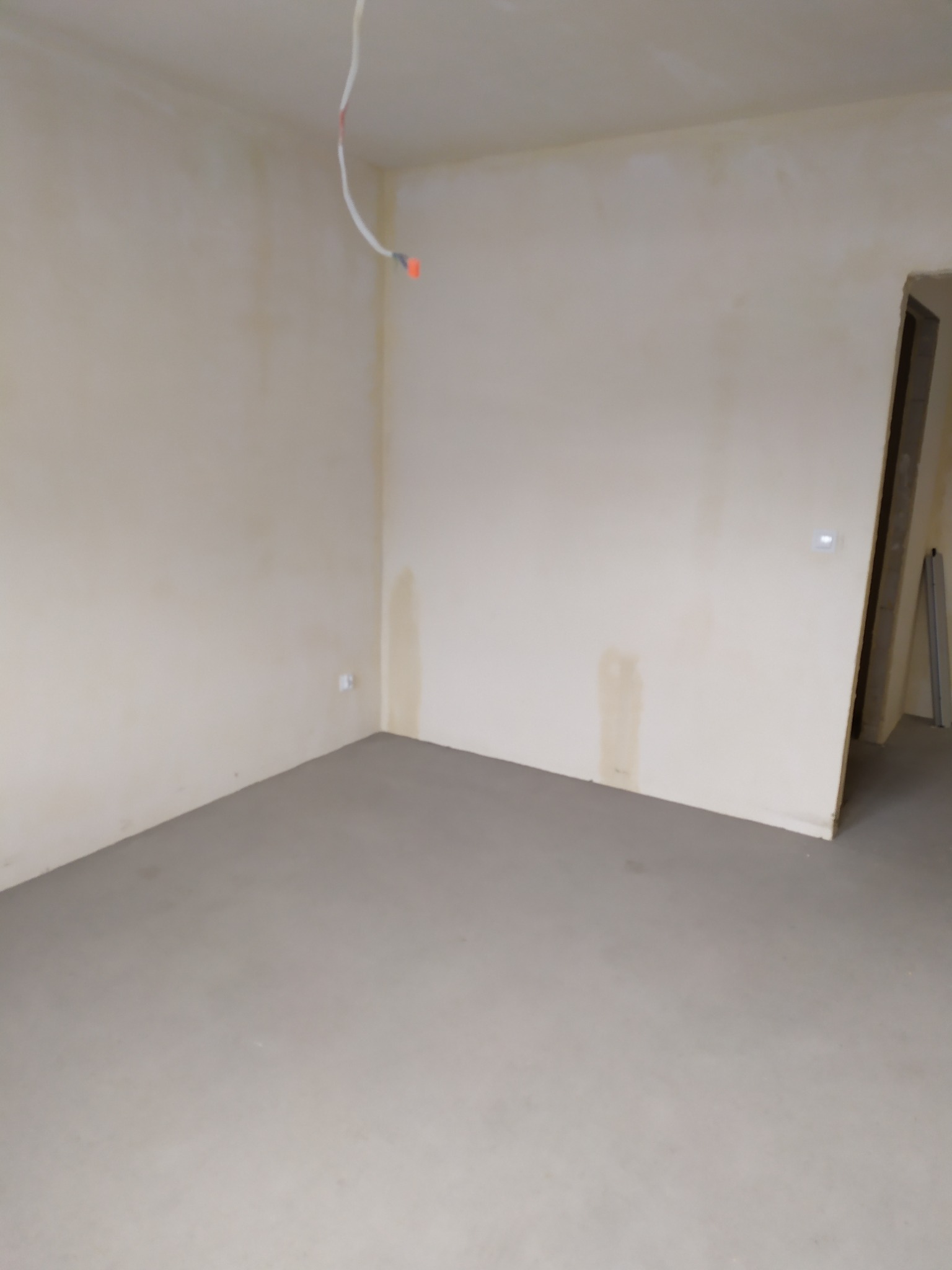 Pusty pokój w trakcie prac wykończeniowych z widocznymi śladami po zalaniach na ścianach, surowy przewód elektryczny zwisający z sufitu i widoczne gniazdko elektryczne na ścianie.