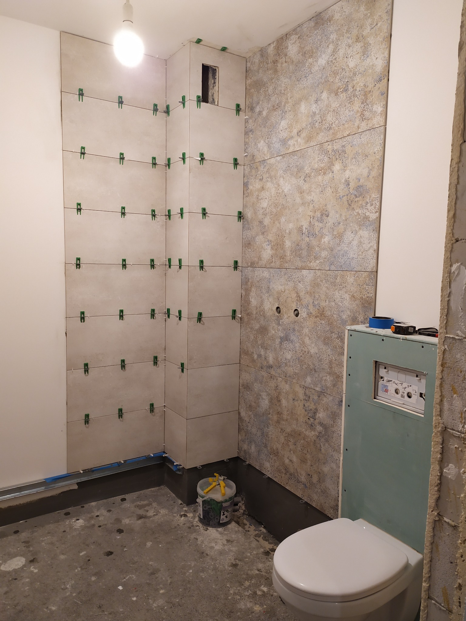 Układanie płytek w łazience, widoczne płytki z zielonymi klipsami poziomującymi, częściowo ułożona ściana z płytek o wzorzystej powierzchni, stelaż podtynkowy WC zabudowany płytą gipsową, surowy...