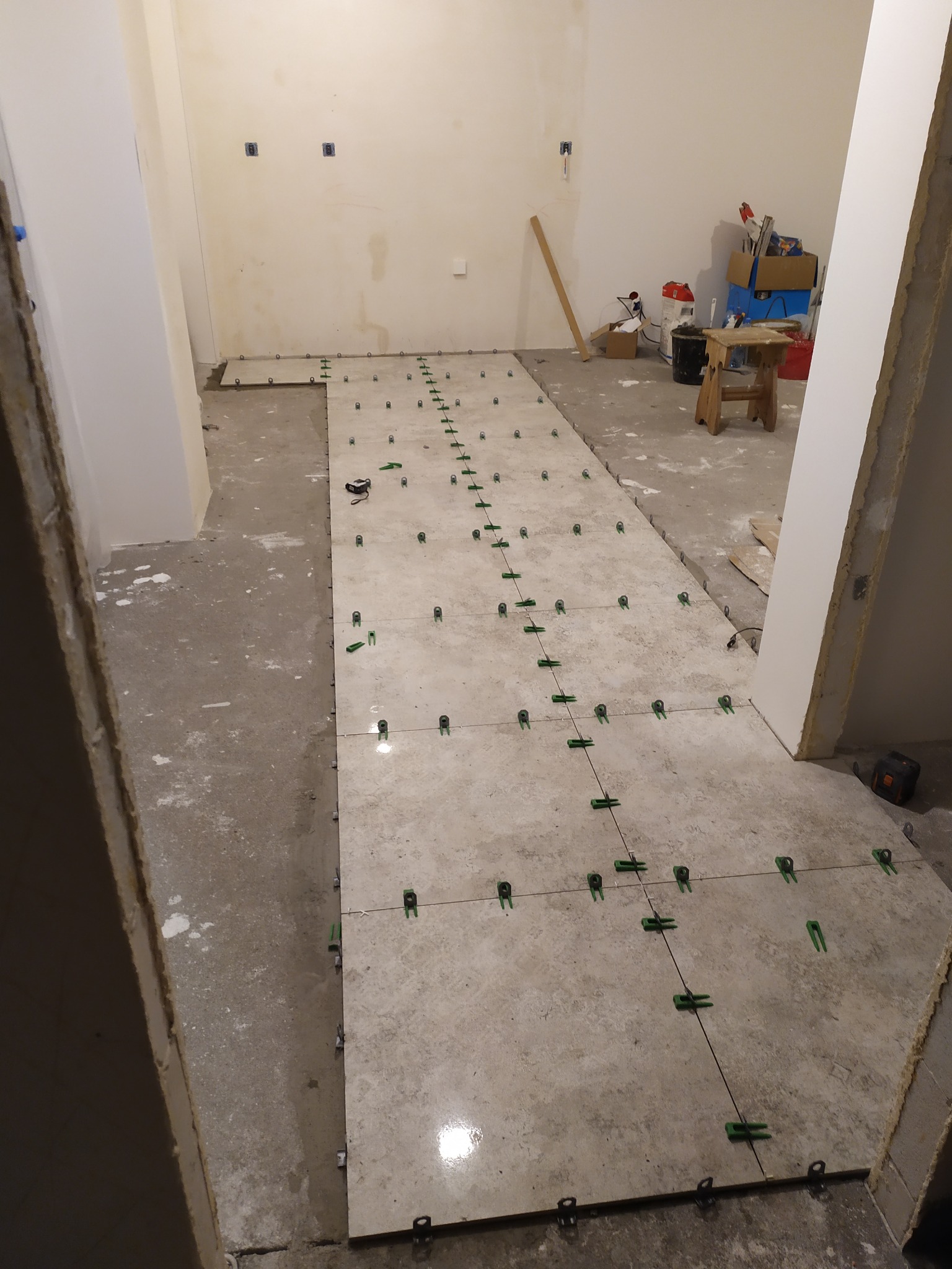 Układanie jasnych płytek podłogowych z użyciem systemu poziomowania, widoczne zielone klipsy i kliny, surowy stan pomieszczenia z widocznymi śladami prac budowlanych.