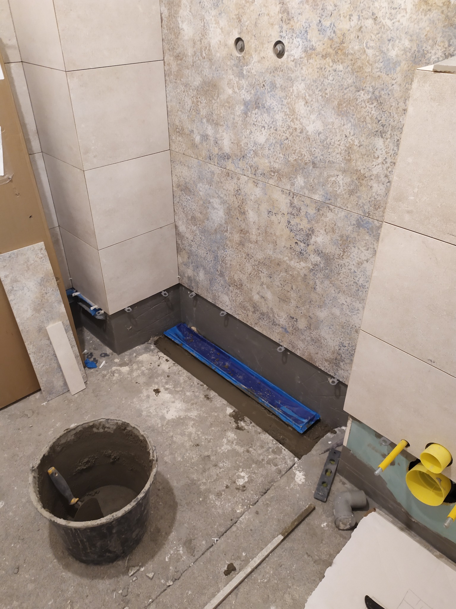 Fragment łazienki w trakcie układania płytek, widoczne narzędzia, wiadro z zaprawą, fragment instalacji wodnej i kanalizacyjnej, oraz płytki o różnej fakturze na ścianach.