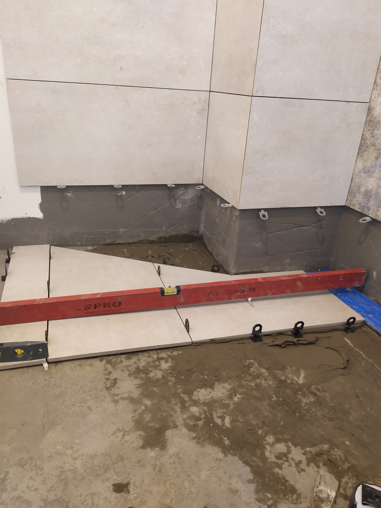 Układanie jasnych płytek podłogowych, z użyciem poziomicy i systemu poziomowania, widoczny świeży klej i elementy montażowe.