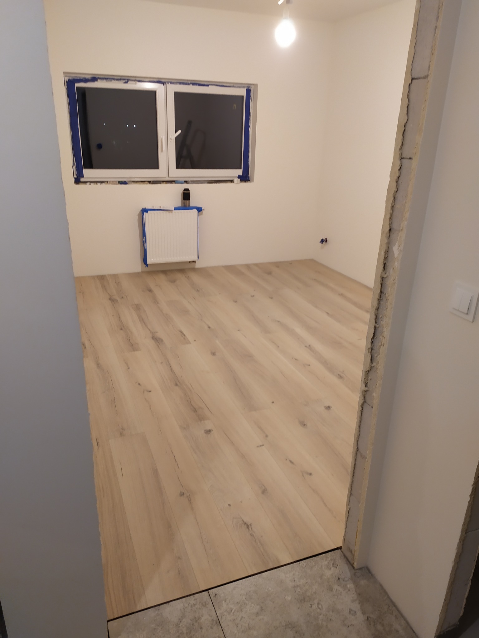 Wnętrze łazienki w trakcie prac wykończeniowych: jasna podłoga imitująca drewno, okno zabezpieczone taśmą malarską, grzejnik z niebieskim zabezpieczeniem, surowe wykończenie ościeżnicy.