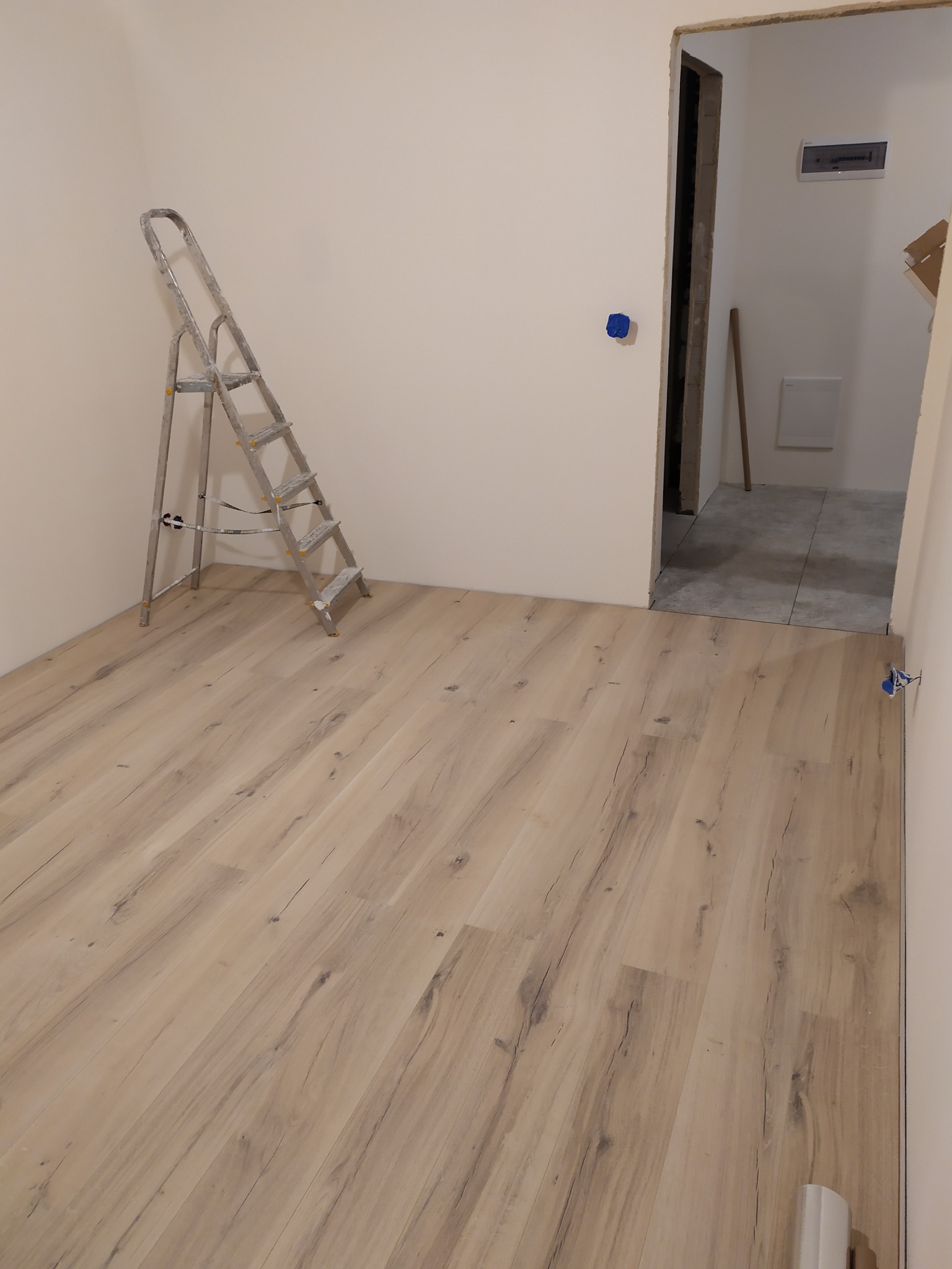 Pusty pokój po remoncie z nową podłogą z paneli, drabina malarska oparta o ścianę, widoczne gniazdko elektryczne i surowa framuga drzwi.