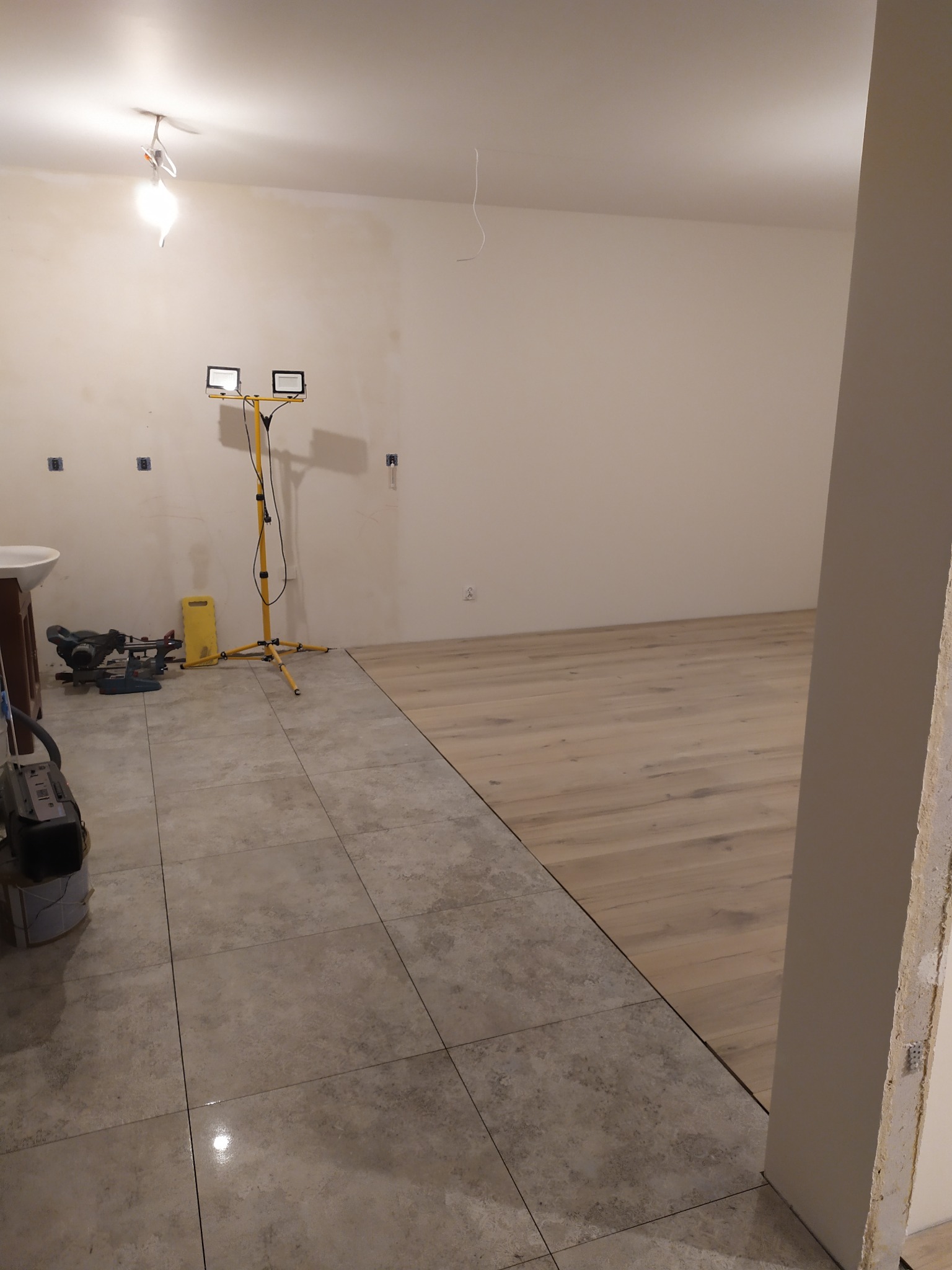 Podłoga w trakcie łączenia dwóch rodzajów wykończenia: płytki ceramiczne i panele drewnopodobne, widoczna lampa budowlana, narzędzia i umywalka.