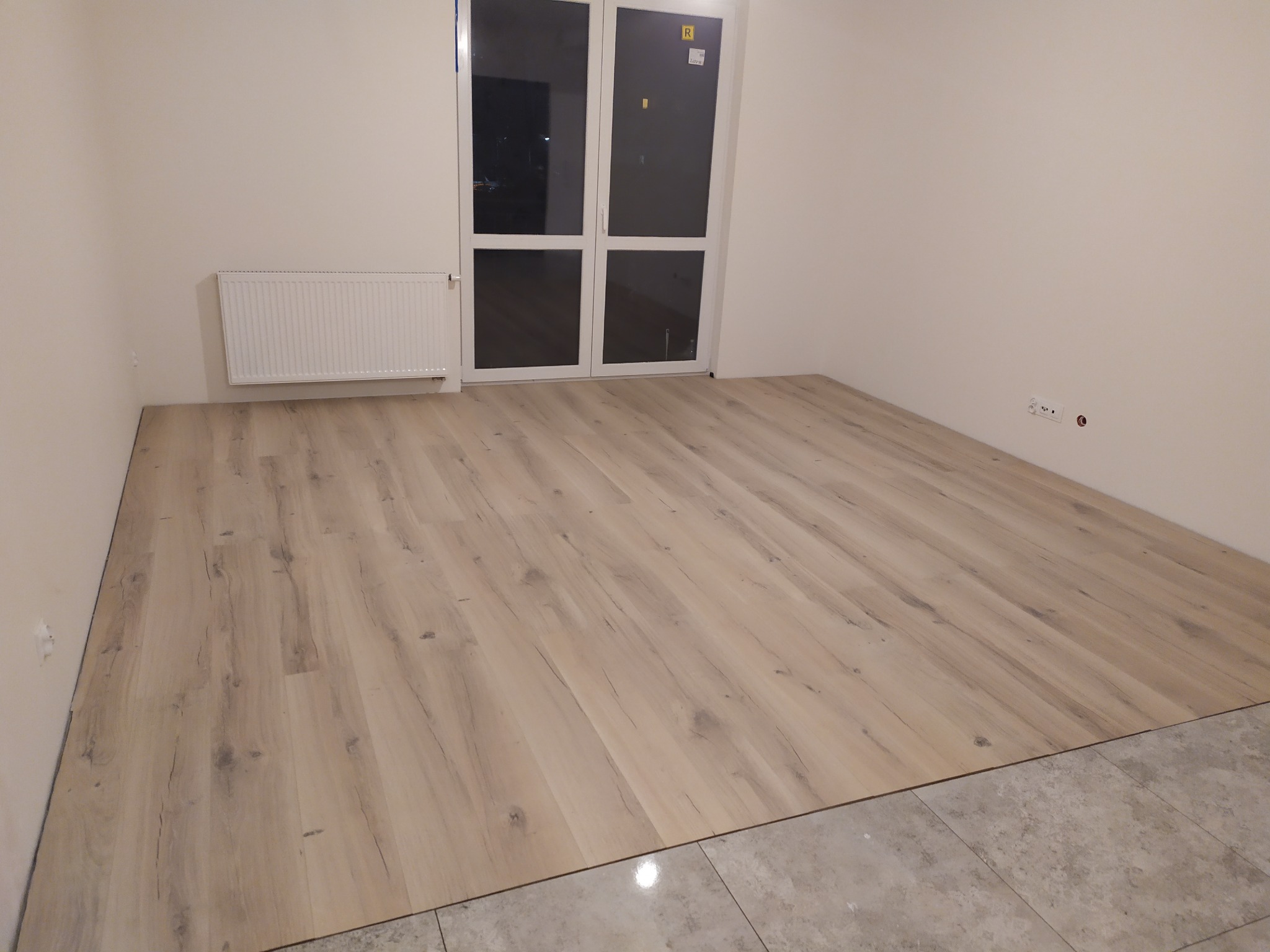 Podłoga z paneli imitujących drewno w jasnym odcieniu, częściowo ułożona na nowym podłożu, widoczne przygotowanie do dalszych prac wykończeniowych w pomieszczeniu z grzejnikiem i podwójnymi...