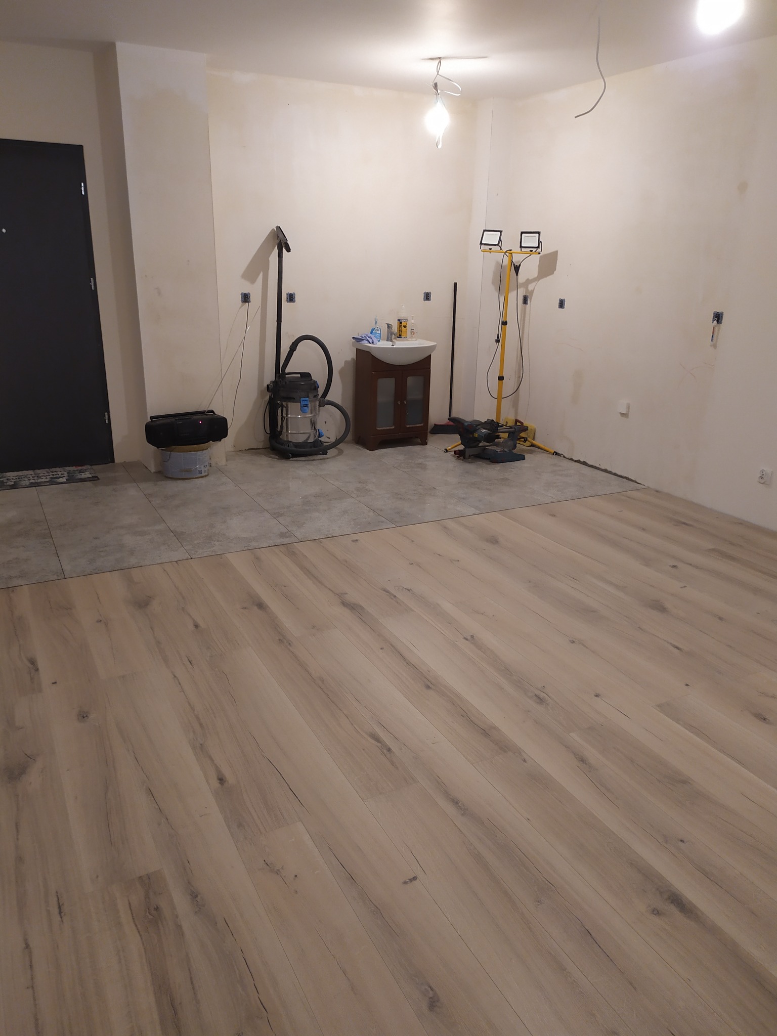 Pokój w trakcie prac wykończeniowych: położona podłoga z paneli, częściowo ułożone płytki, ściany przygotowane pod malowanie, widoczny sprzęt budowlany i narzędzia.