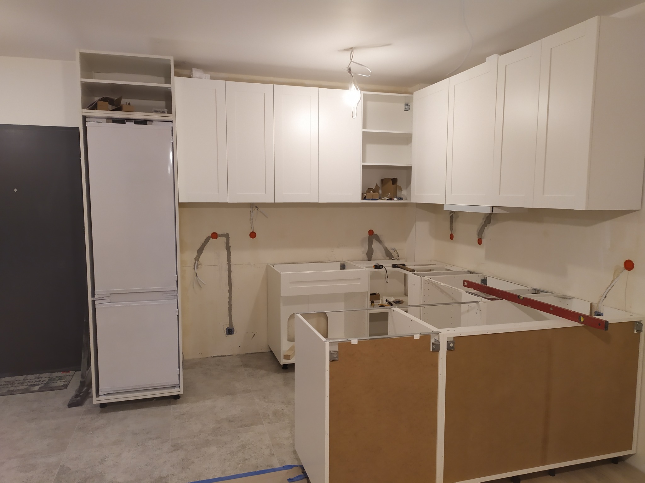 Montaż szafek kuchennych w surowym stanie, widoczne niezabudowane elementy, poziomica na blacie, przygotowane instalacje elektryczne, białe fronty szafek.