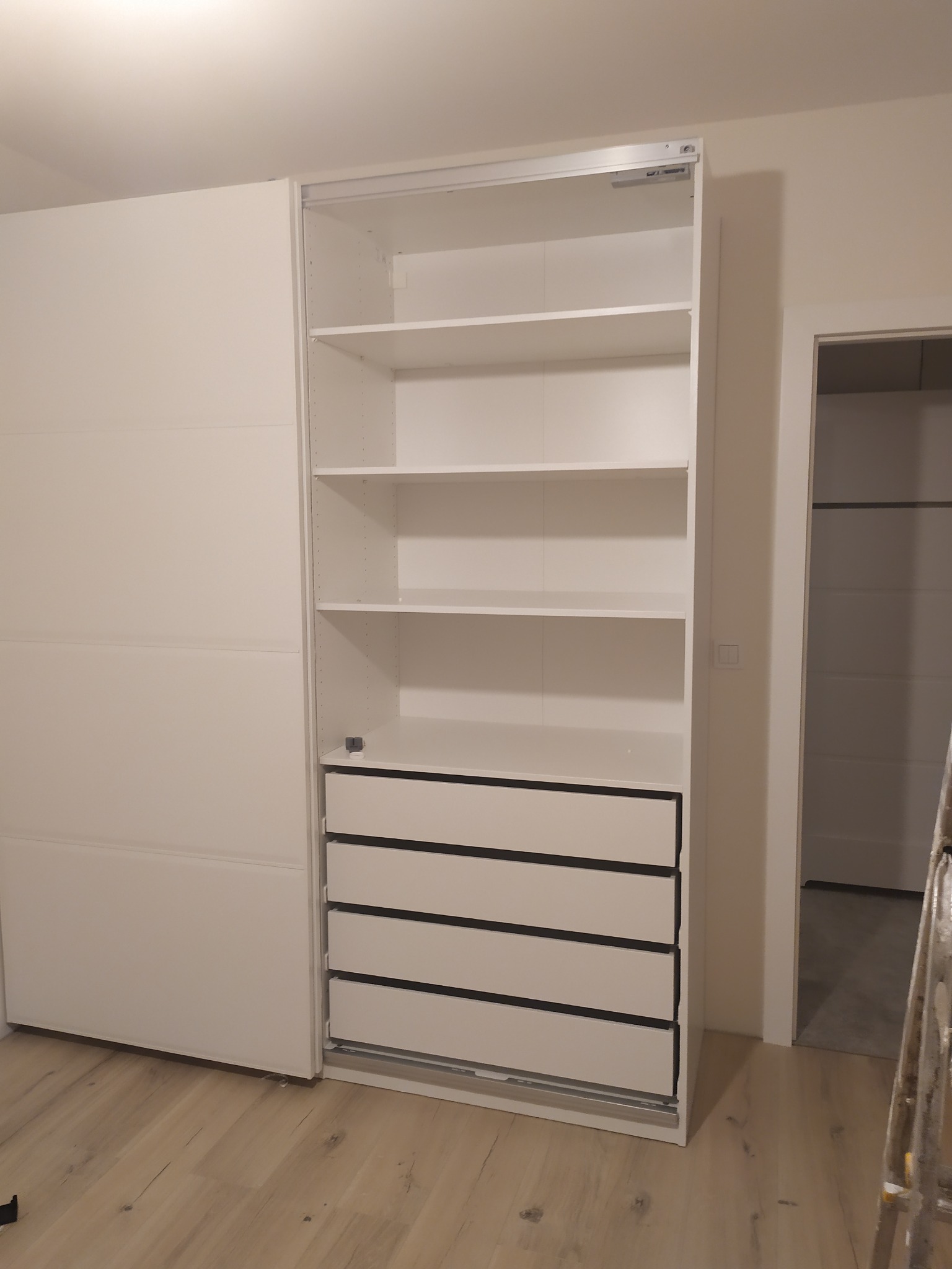 Biała, częściowo otwarta szafa wnękowa z przesuwnymi drzwiami i szufladami, widoczne półki, montaż w trakcie wykończenia wnętrza, drabina oparta o ścianę w tle.
