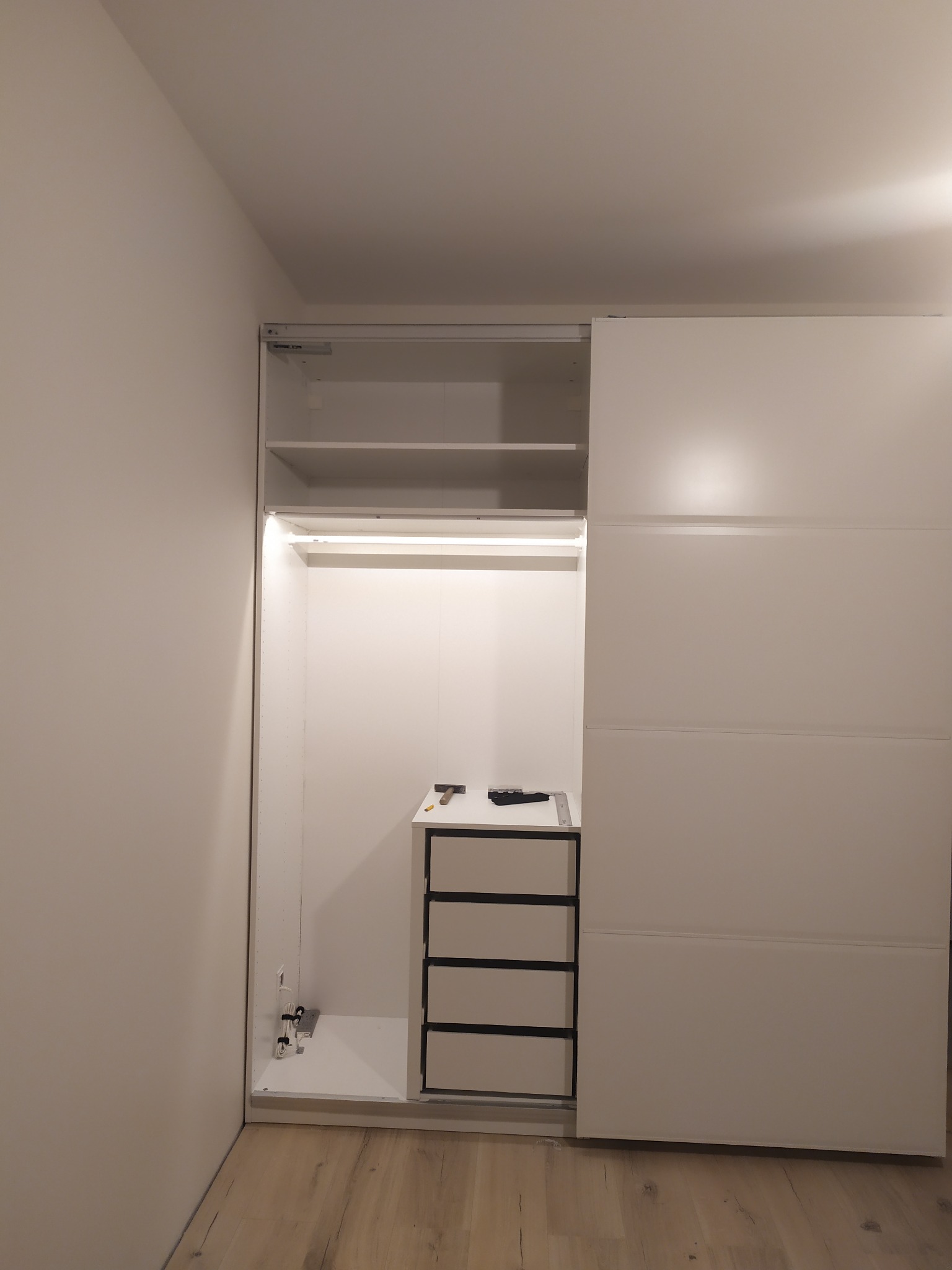 Wnętrze szafy wnękowej z drzwiami przesuwnymi w trakcie montażu, widoczne półki, szuflady, oświetlenie LED oraz narzędzia na blacie.