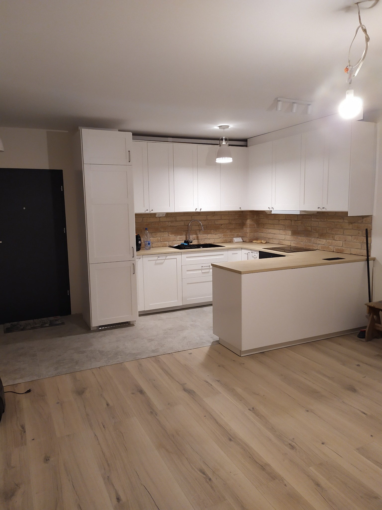 Nowoczesna, jasna kuchnia z białymi szafkami, ceglaną ścianą nad blatem i drewnianą podłogą. Widoczny fragment wyspy kuchennej z drewnianym blatem oraz drzwi wejściowe po lewej stronie.