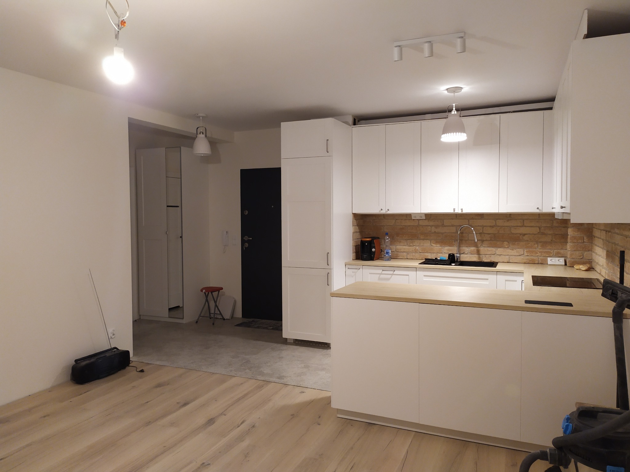 Wnętrze przestronnej kuchni z białymi szafkami, drewnianym blatem i ceglaną ścianą nad blatem, podłoga z jasnych paneli, widoczny odkurzacz i radio, pomieszczenie w trakcie wykańczania.