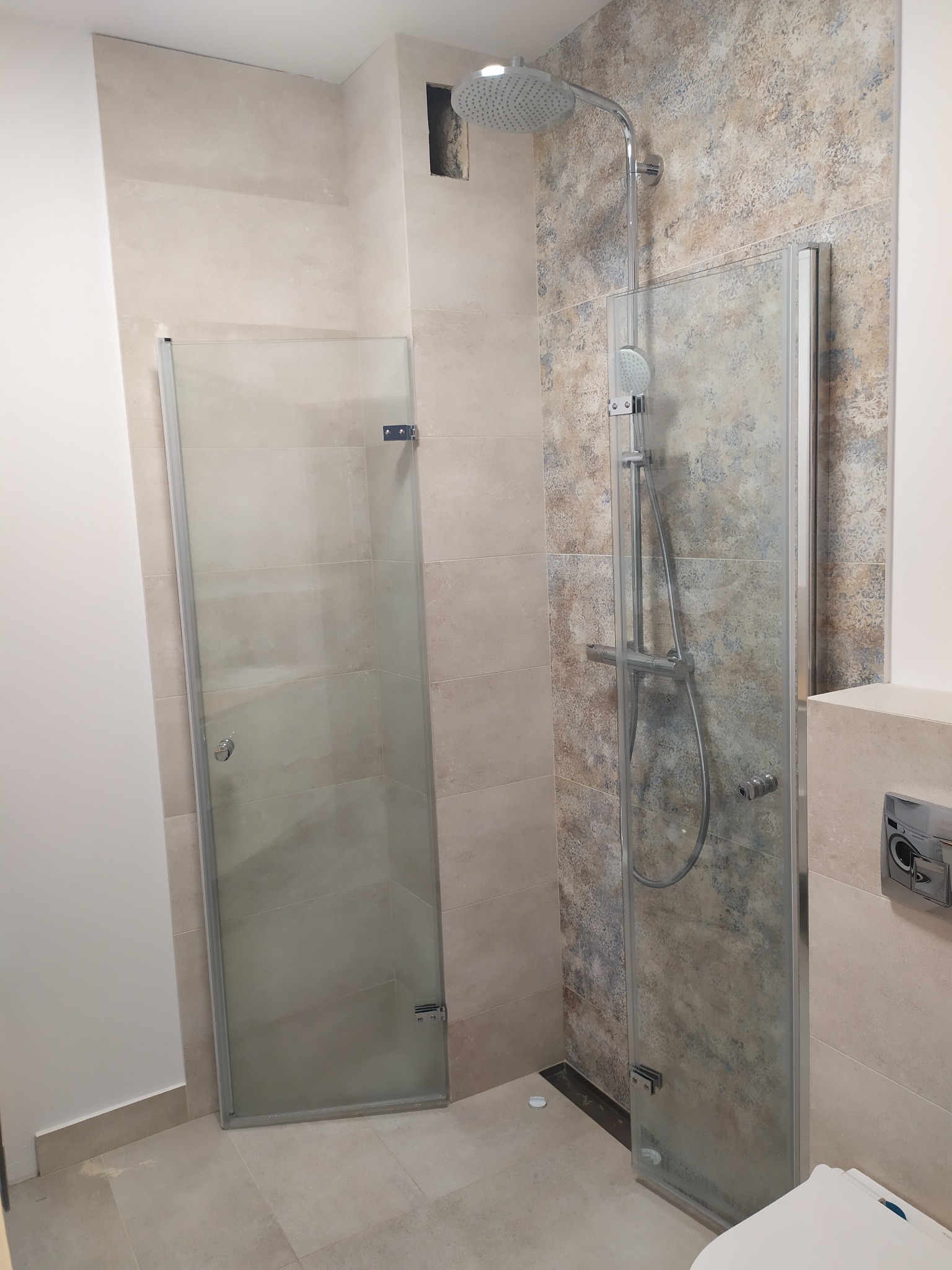 Nowoczesna kabina prysznicowa z dwoma szklanymi ściankami, chromowana armatura i deszczownica na tle beżowych i wzorzystych płytek w łazience.