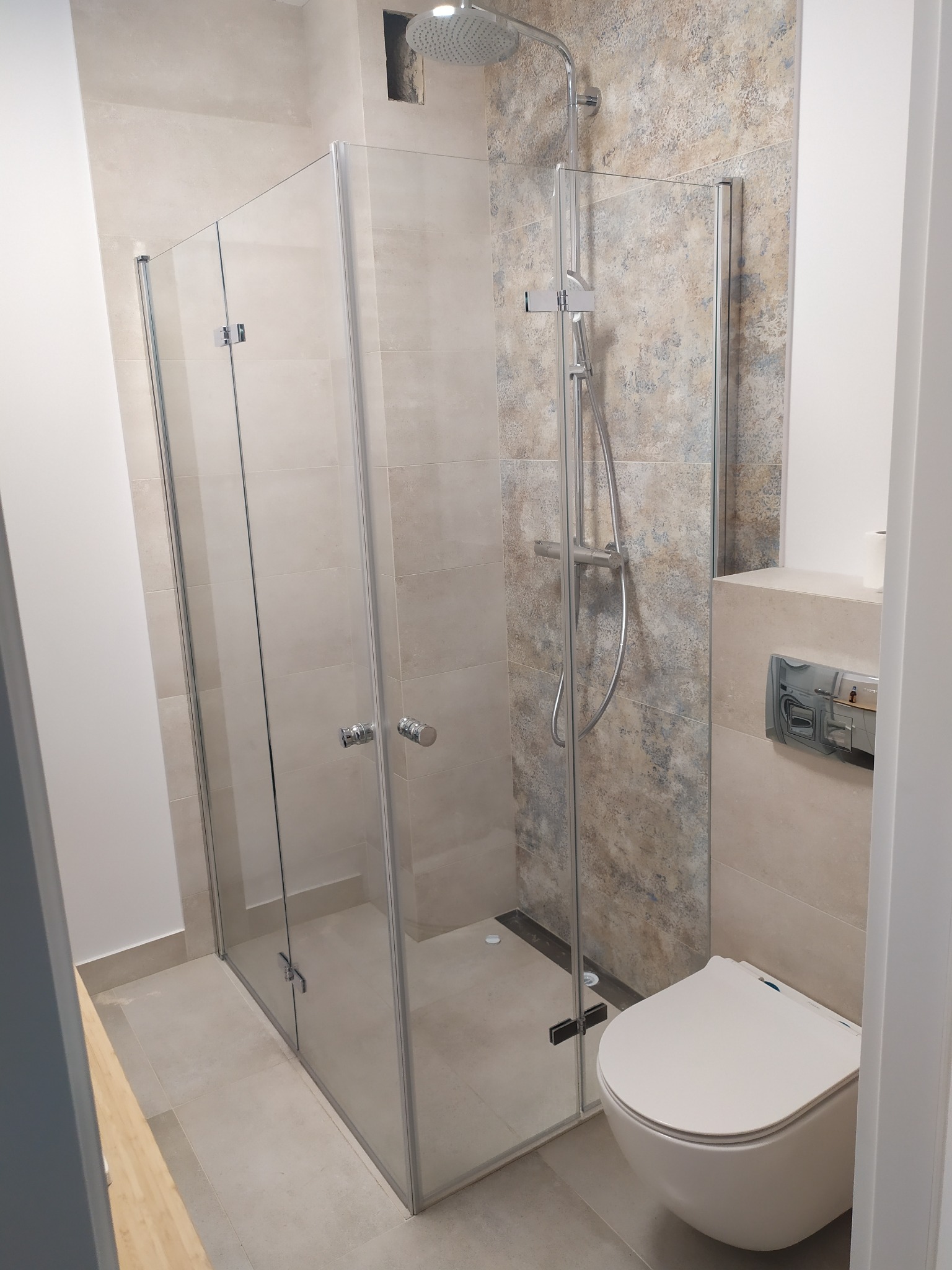 Nowoczesna łazienka z kabiną prysznicową typu walk-in, wiszącą toaletą i beżowymi płytkami imitującymi kamień na ścianach i podłodze, widoczne chromowane elementy armatury.