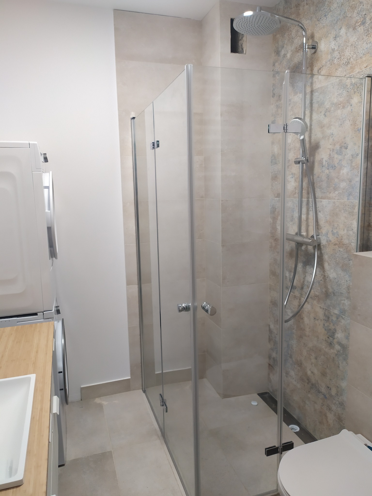 Nowoczesna kabina prysznicowa ze szklanymi drzwiami, deszczownicą i odpływem liniowym w wykończonej łazience z pralką i umywalką w tle.