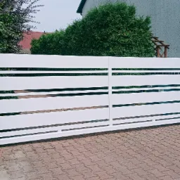 Nowoczesna, biała brama wjazdowa przesuwna z poziomymi panelami, osadzona między ceglanymi słupkami na tle kostki brukowej i zieleni.