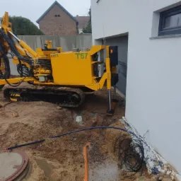 Żółta wiertnica Beretta T57 na gąsienicach, przygotowana do wiercenia przy białym budynku z szarą bramą garażową, widoczne kable i rury w wykopie.