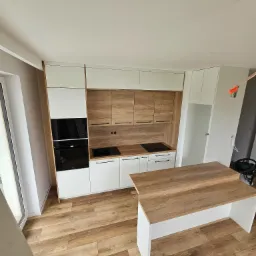 Nowoczesna kuchnia z drewnianymi akcentami, widok z góry na zabudowę z piekarnikiem, płytą indukcyjną i wyspą kuchenną z blatem imitującym drewno, na podłodze panele podłogowe imitujące drewno...