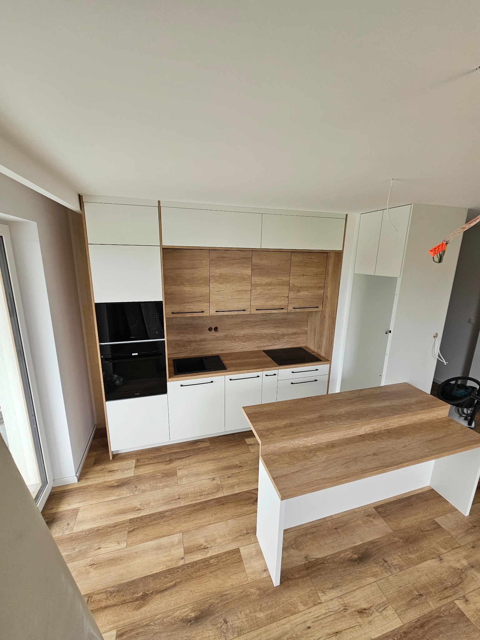 Nowoczesna kuchnia z drewnianymi akcentami, widok z góry na zabudowę z piekarnikiem, płytą indukcyjną i wyspą kuchenną z blatem imitującym drewno, na podłodze panele podłogowe imitujące drewno...