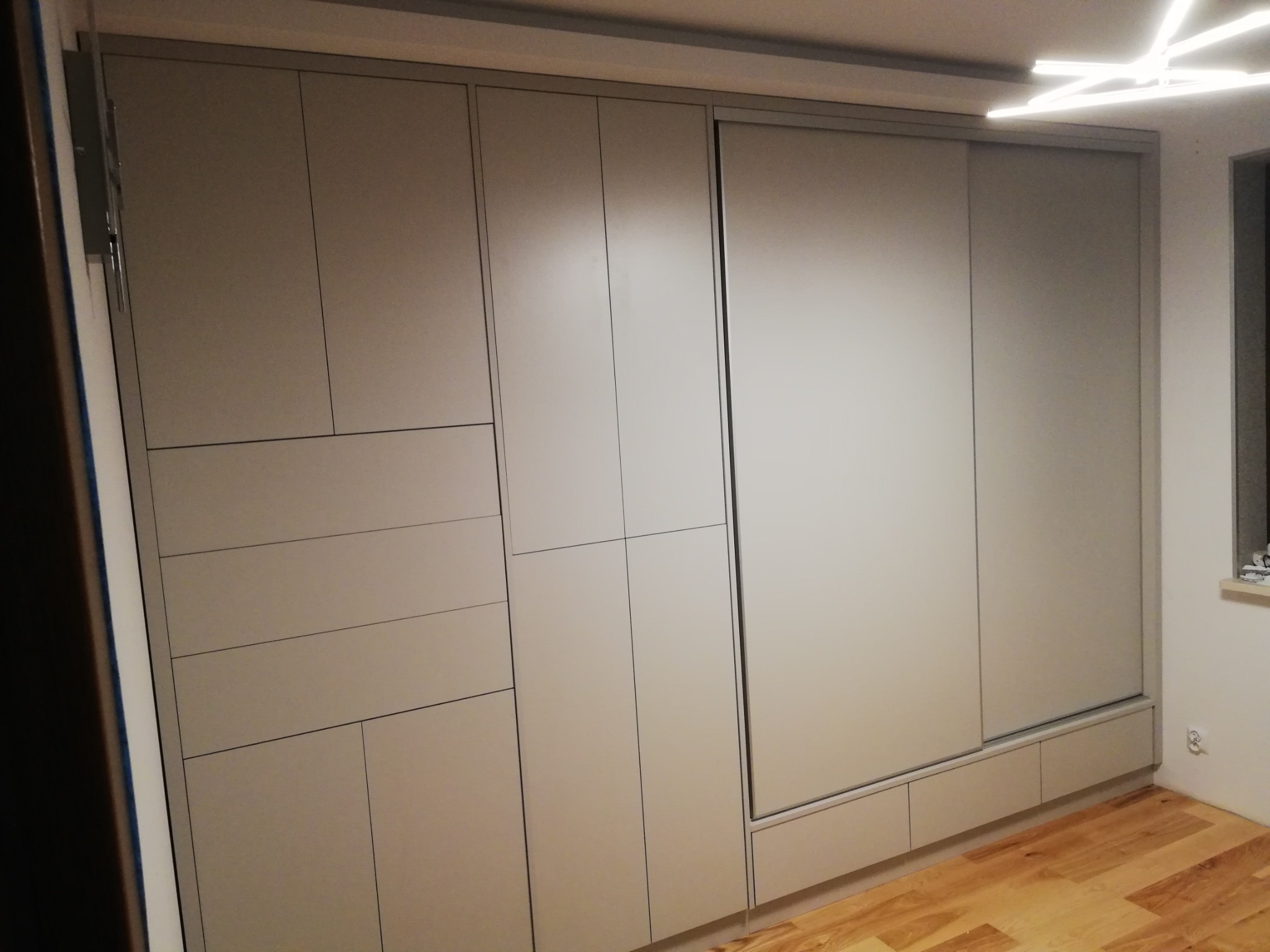 Jasnoszara szafa wnękowa z przesuwnymi drzwiami i geometrycznym układem frontów, widoczna z boku w nowym mieszkaniu z parkietem i nowoczesnym oświetleniem sufitowym.