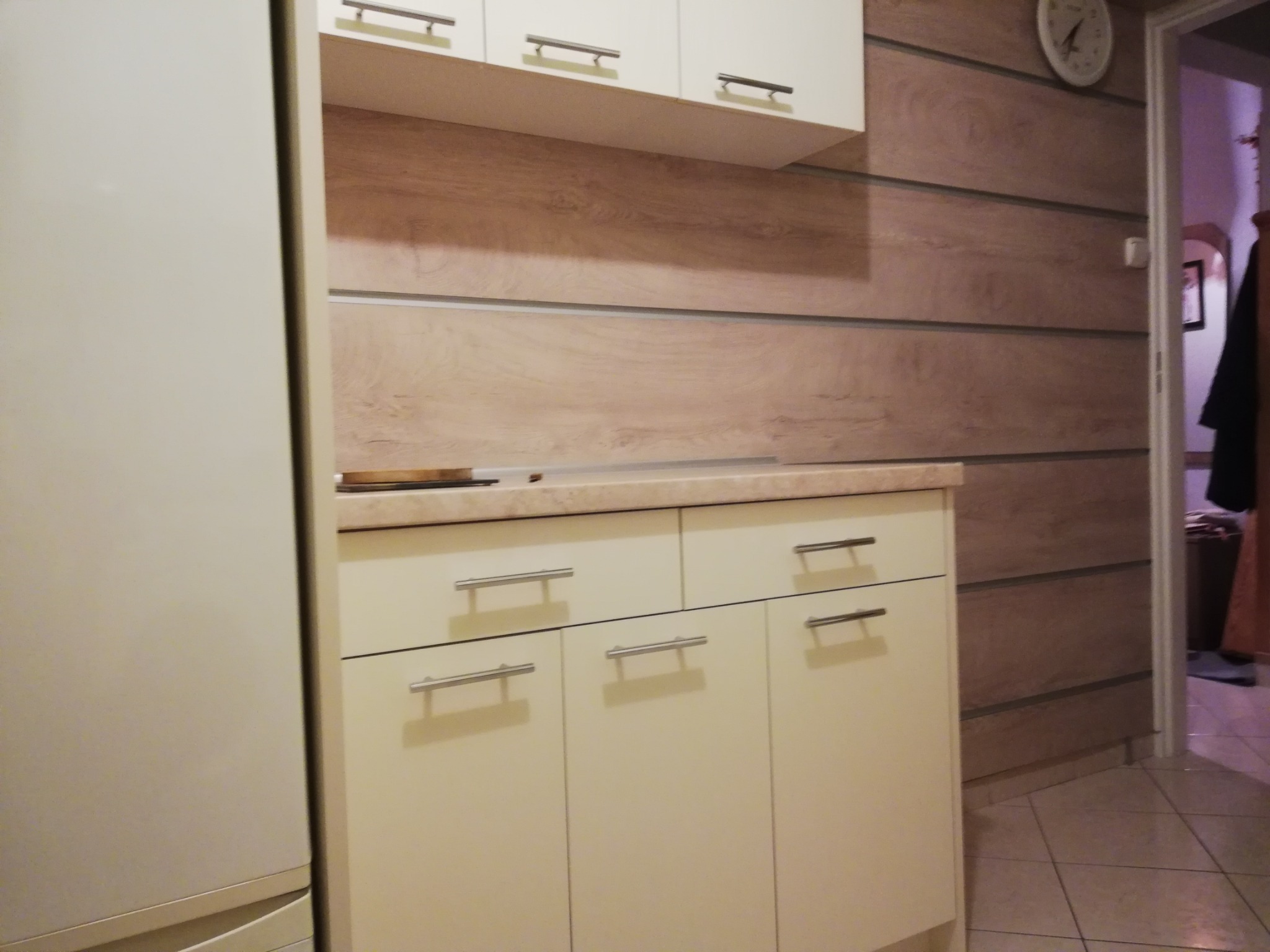 Beżowa zabudowa kuchenna z szafkami wiszącymi i stojącymi, metalowe uchwyty, ściana wykończona panelami imitującymi drewno, widoczny fragment lodówki.