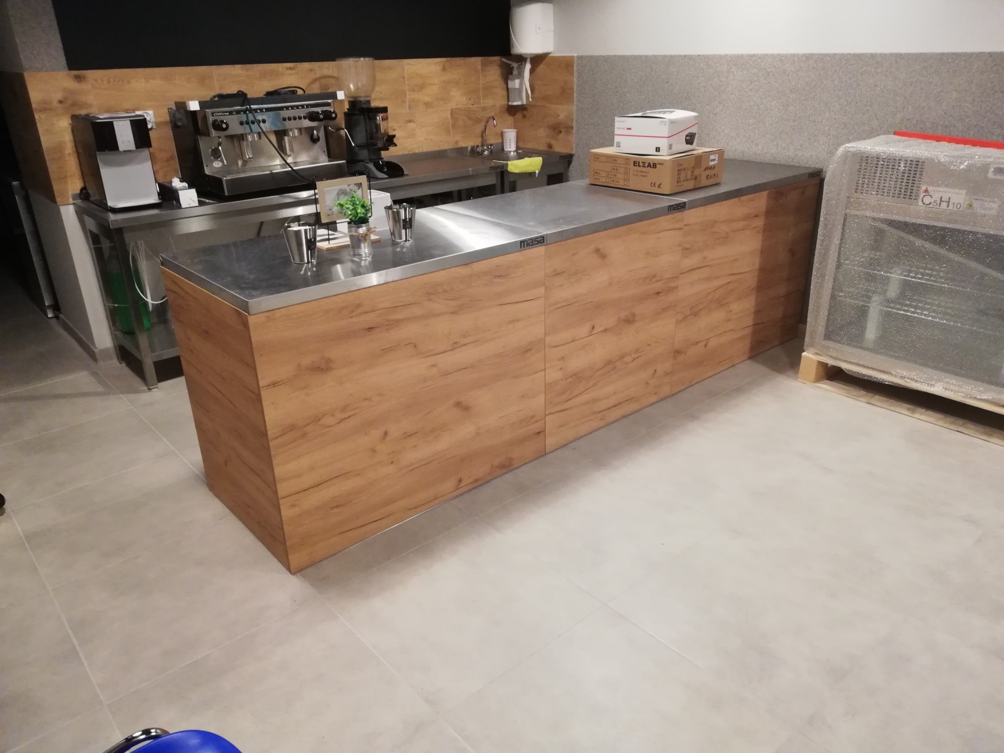 Drewniana lada z metalowym blatem w lokalu gastronomicznym, z ekspresami do kawy i zlewem w tle, oraz zapakowaną chłodziarką po prawej stronie.