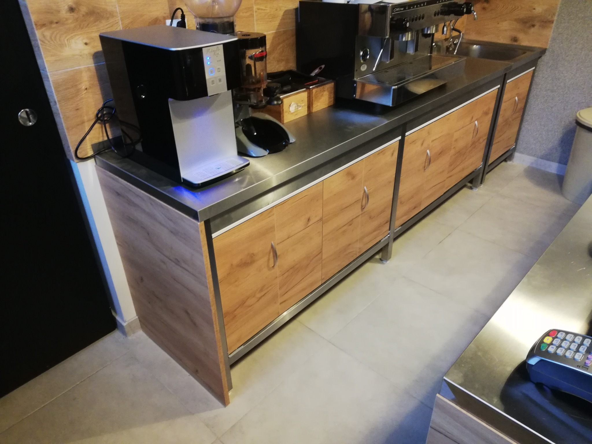 Długa, drewnopodobna szafka kuchenna z metalowym blatem, na którym stoją ekspres do kawy, dystrybutor wody i akcesoria.