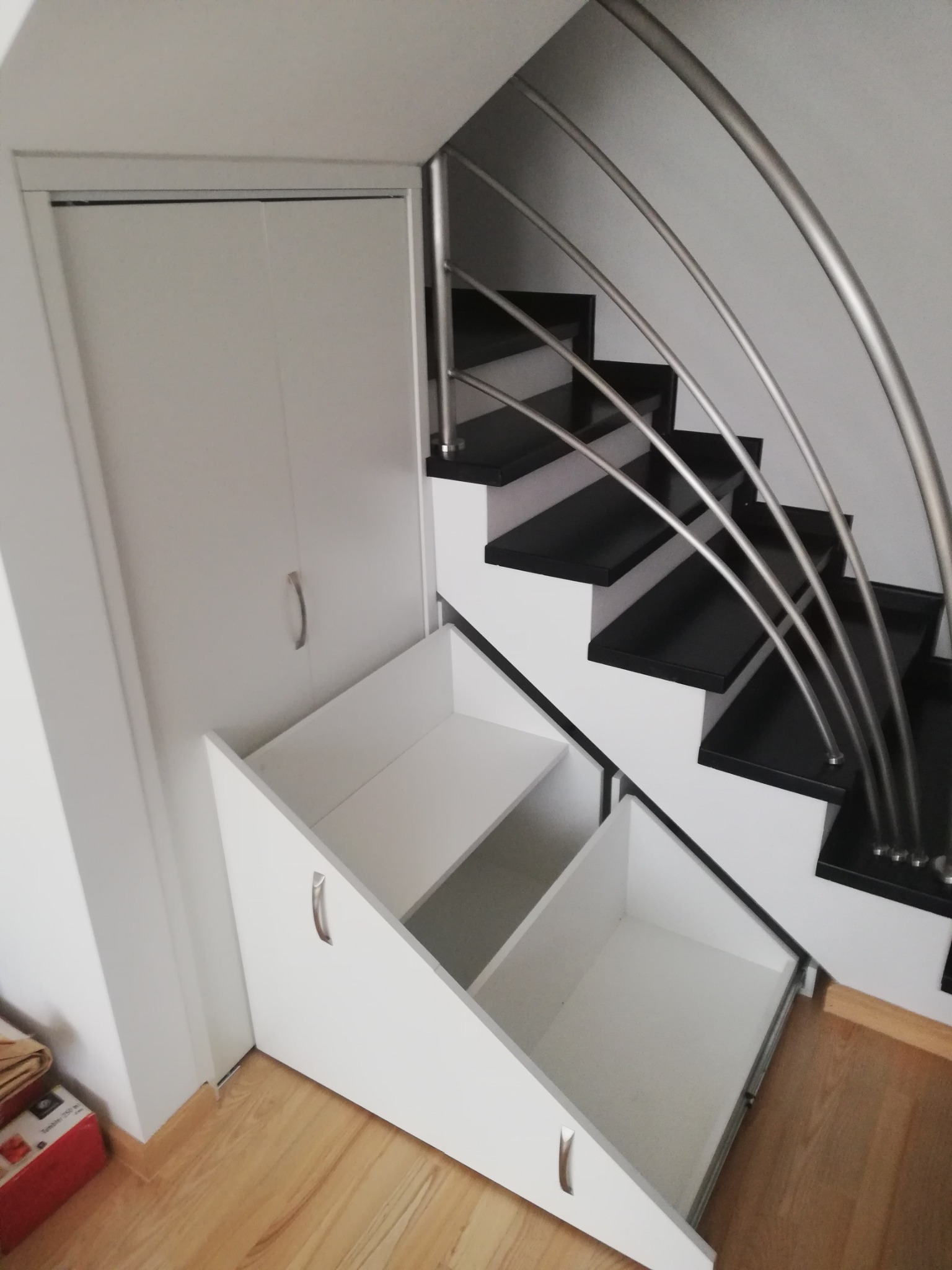 Biała szafa wnękowa pod schodami z wysuwanymi szufladami, czarne schody z metalową balustradą, widok z góry na drewnianą podłogę.