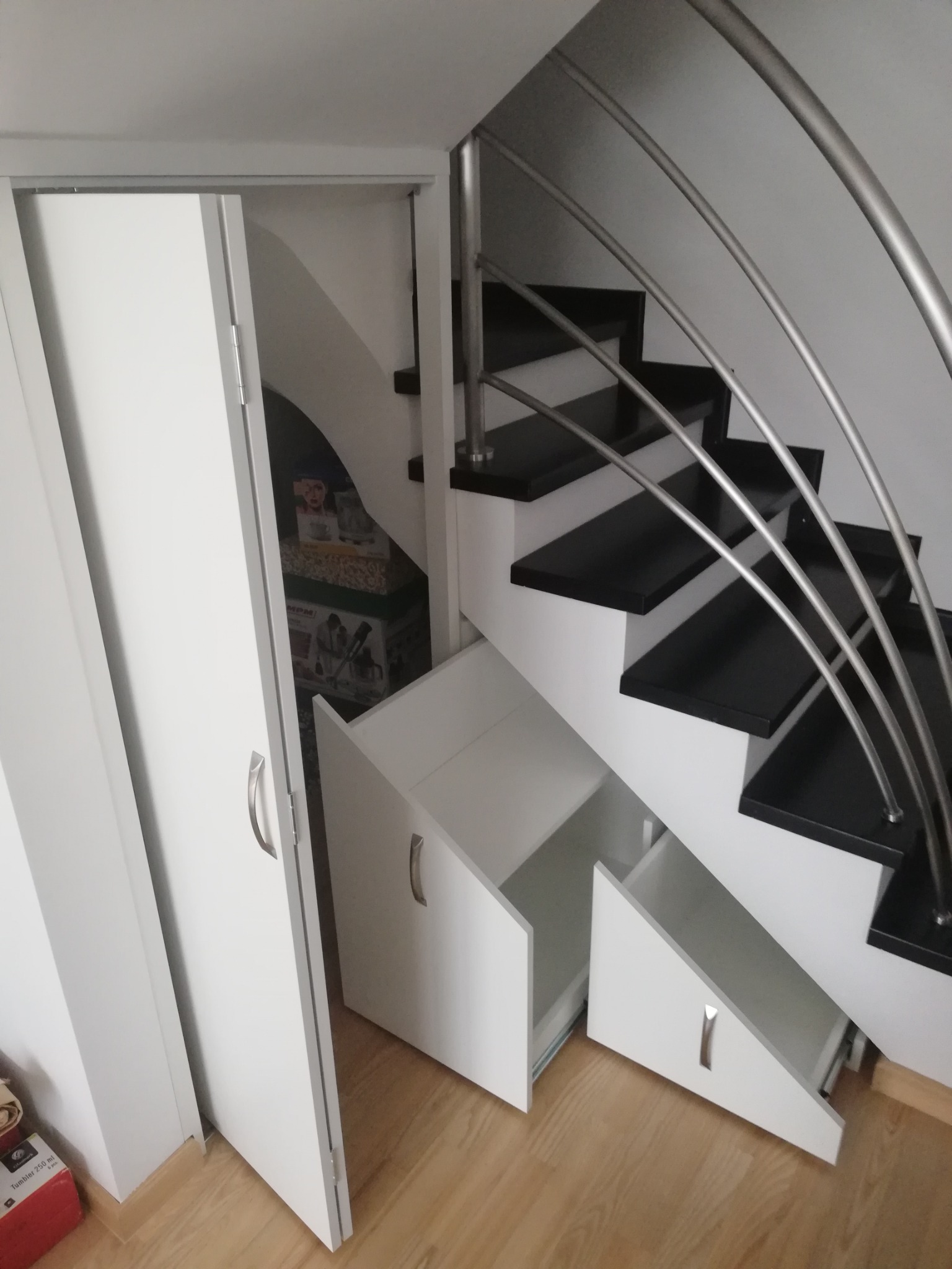 Biała szafa przesuwna i szuflady pod schodami z czarnymi stopniami i srebrną balustradą, otwarte szuflady ukazują puste wnętrze, w szafie widoczne pudełka z urządzeniami AGD.