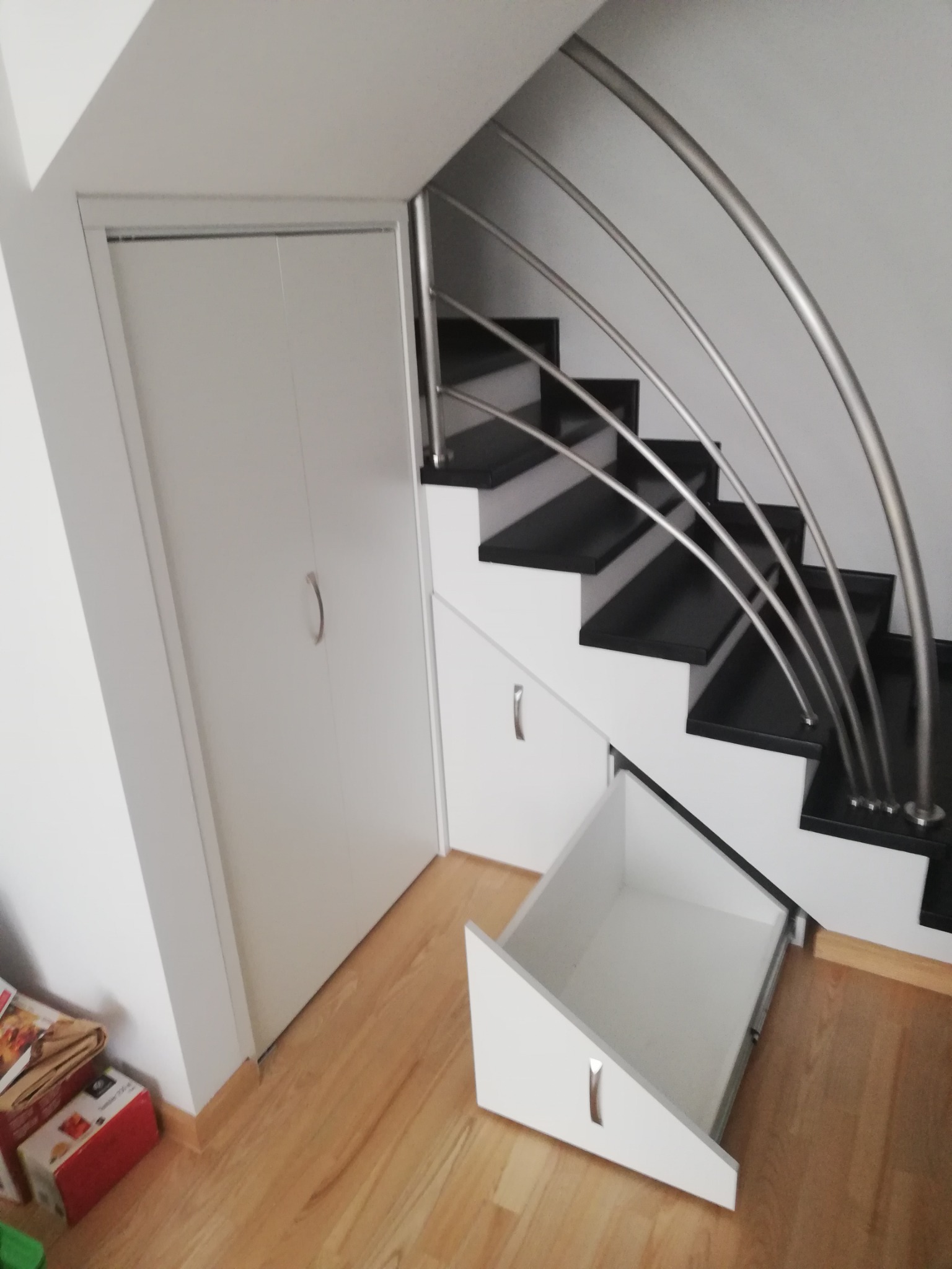 Biała szafa wnękowa z drzwiami przesuwnymi obok schodów z czarnymi stopniami i metalową balustradą; pod schodami wysuwana szuflada z białym frontem.