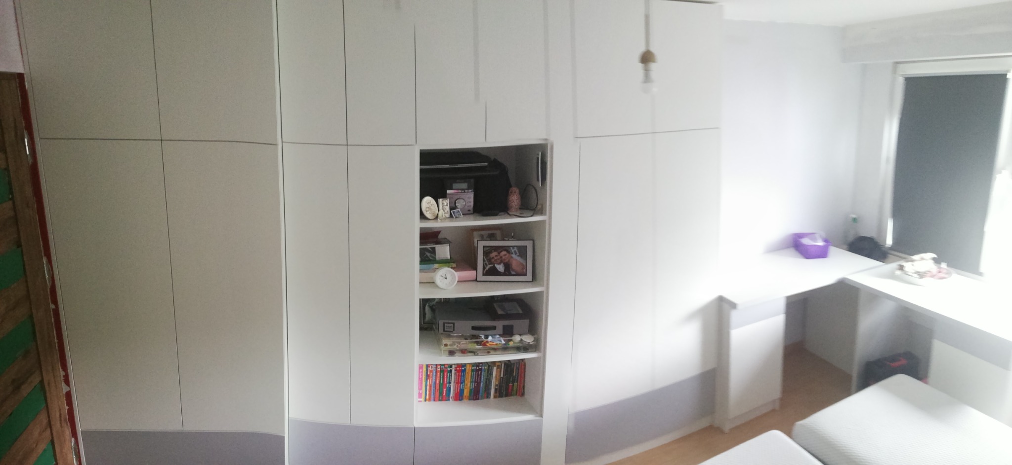 Biała szafa wnękowa z półkami, na których stoją książki, ramki ze zdjęciami i dekoracje, uzupełniona o biurko w podobnej stylistyce.